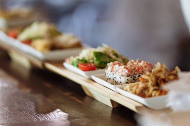 Masanın arka planında ahşap standlar üzerinde beyaz kare tabaklarda çeşitli Japon yemekleri