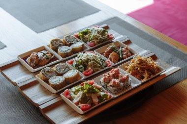 Masanın arka planında ahşap standlar üzerinde beyaz kare tabaklarda çeşitli Japon yemekleri