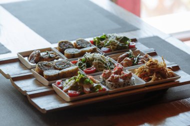 Masanın arka planında ahşap standlar üzerinde beyaz kare tabaklarda çeşitli Japon yemekleri