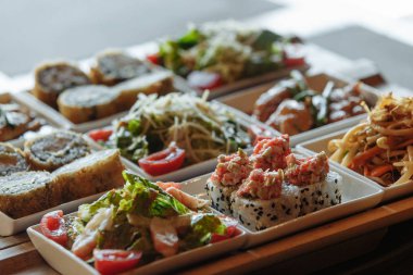 Masanın arka planında ahşap standlar üzerinde beyaz kare tabaklarda çeşitli Japon yemekleri