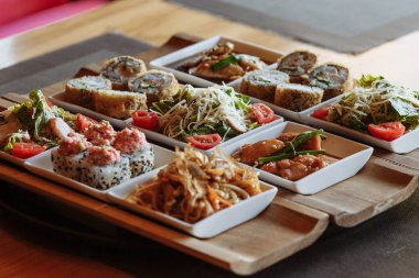 Masanın arka planında ahşap standlar üzerinde beyaz kare tabaklarda çeşitli Japon yemekleri