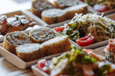 Masanın arka planında ahşap standlar üzerinde beyaz kare tabaklarda çeşitli Japon yemekleri