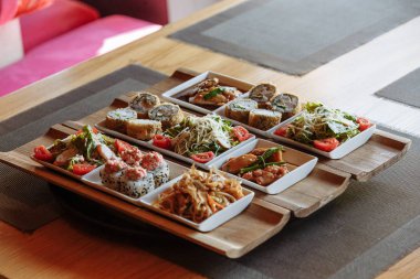 Masanın arka planında ahşap standlar üzerinde beyaz kare tabaklarda çeşitli Japon yemekleri