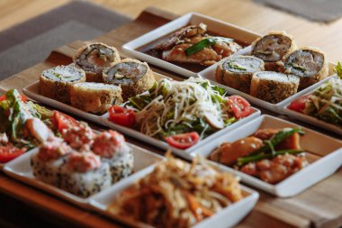Masanın arka planında ahşap standlar üzerinde beyaz kare tabaklarda çeşitli Japon yemekleri