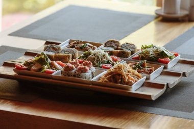 Masanın arka planında ahşap standlar üzerinde beyaz kare tabaklarda çeşitli Japon yemekleri