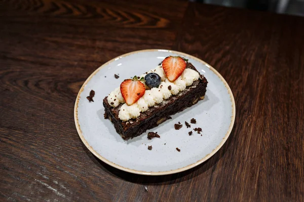Brownie adornado con crema batida, fresas y arándanos en un plato sobre un fondo de madera oscura — Foto de Stock