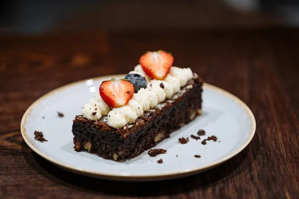 Brownie adornado con crema batida, fresas y arándanos en un plato sobre un fondo de madera oscura — Foto de Stock