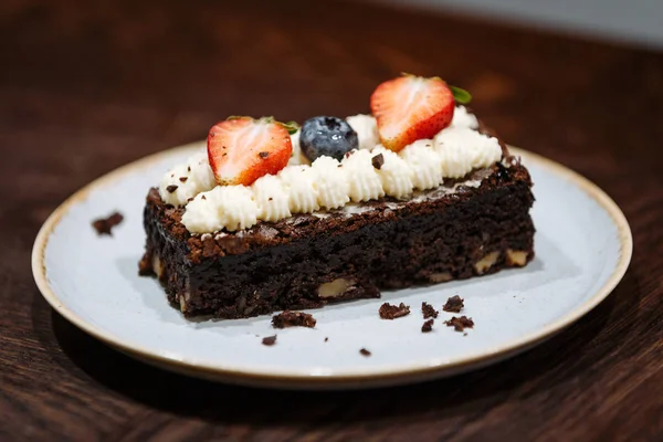 Brownie adornado con crema batida, fresas y arándanos en un plato sobre un fondo de madera oscura — Foto de Stock