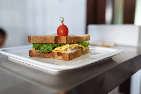 sandwich with cheese and greens decorated with tomato in a white rectangular plate on a metal table
