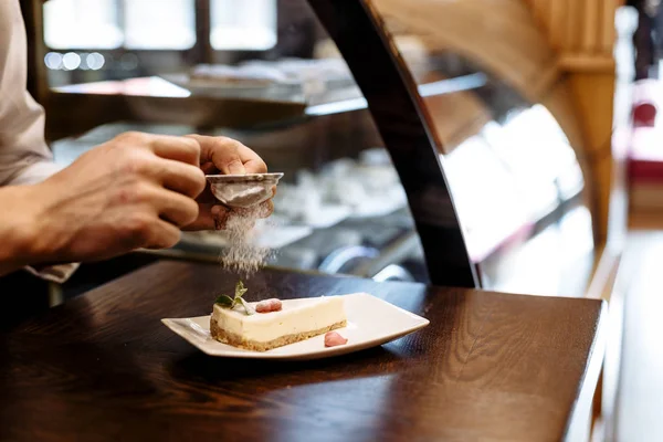 Chef espolvoreado con el tamiz de azúcar en polvo de un pedazo de pastel en un plato sobre una mesa de madera — Foto de Stock