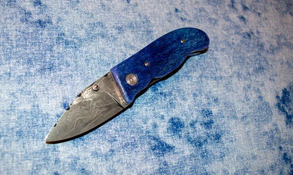Damascus pocket knife with blue camel bone handle displayed on blue and white fabric.