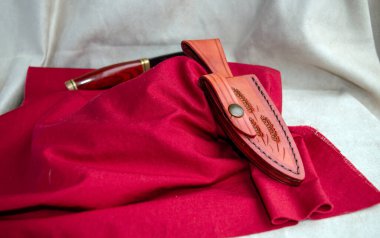 Benzersiz dekore edilmiş bir deri kılıf kırmızı bir kumaş üzerinde sergilenirken, bıçak arka planda, kılıfına dikkat çekmek için bokeh efektiyle oturur..