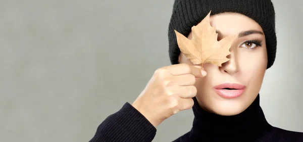 Autumn concept. Beauty portrait of a young woman in warm polo neck top and beanie holding a dried maple leaf to her eye with a sensual look over a grey background with copyspace