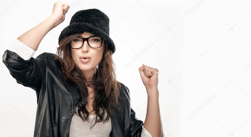 Woman celebrating victory. Chic young woman with long brunette hair wearing modern dark framed glasses and black hat celebrating with raised fists while pouting her lips at camera. isolated on white