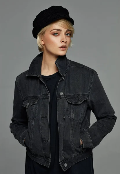 黒いデニムジャケットと灰色の背景にキャップを着て短い髪を持つ若いブロンドの女性の肖像画 — ストック写真