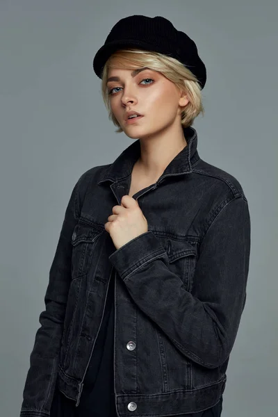 Portrait of young blond woman with short hair wearing black denim jacket and cap on gray background