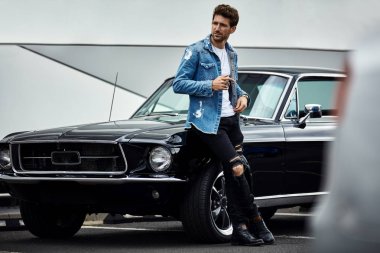 Handsome man in denim jacket posing with black classic car clipart