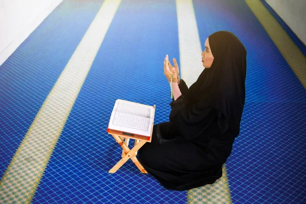 Vista lateral da jovem mulher muçulmana orando a Deus com as mãos para cima na frente do Alcorão Sagrado dentro de uma mesquita — Fotografia de Stock