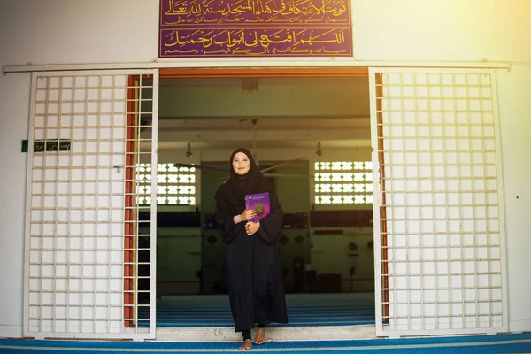 Ung muslimsk kvinna i svart abaya klänning håller Den Heliga Koranen inne i en moské — Stockfoto