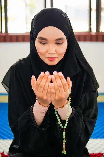 Young muslim woman praying to Allah with hands up on praying mat, holding beads