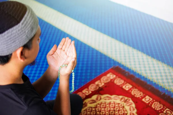 Vista lateral del hombre musulmán haciendo du 'a mientras sostiene cuentas rezando en la alfombra de oración dentro de una mezquita — Foto de Stock