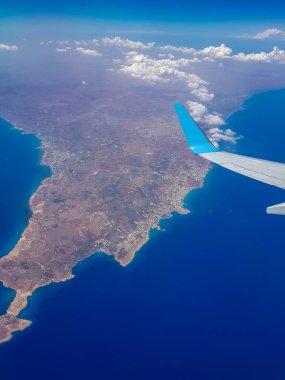 Bir uçağın kanadının altındaki lombozdan Akdeniz adasının manzarası bulutların arasında..