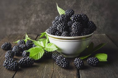 Koyu bir arka plan üzerinde eski ahşap panoları üzerinde bir kase yaprakları ile Olgun böğürtlen.