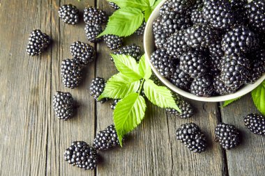 Koyu bir arka plan üzerinde eski ahşap panoları üzerinde bir kase yaprakları ile Olgun böğürtlen.