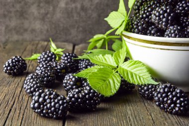 Koyu bir arka plan üzerinde eski ahşap panoları üzerinde bir kase yaprakları ile Olgun böğürtlen.