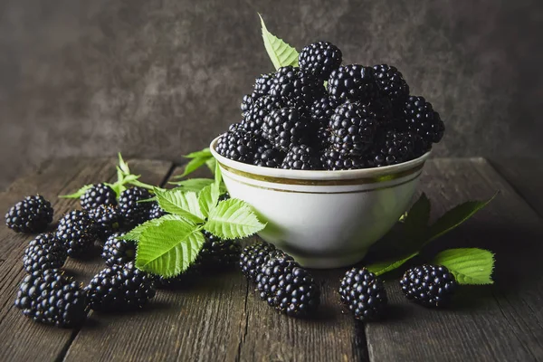 Koyu bir arka plan üzerinde eski ahşap panoları üzerinde bir kase yaprakları ile Olgun böğürtlen.