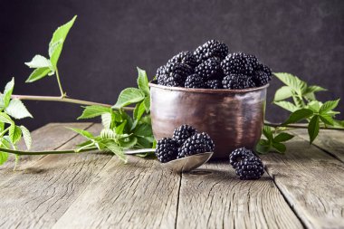 Eski ahşap tahtalar üzerinde eski bakır tabaklarda yaprakları ile Olgun böğürtlen ve koyu bir arka plan üzerinde bir kaşık yanında yalan.