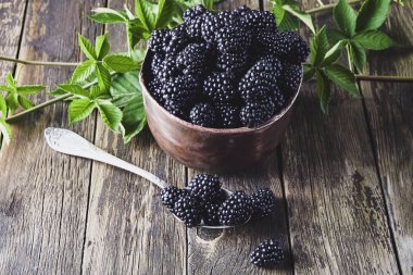 Eski ahşap tahtalar üzerinde eski bakır tabaklarda yaprakları ile Olgun böğürtlen ve koyu bir arka plan üzerinde bir kaşık yanında yalan.