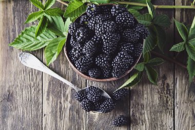 Eski ahşap tahtalar üzerinde eski bakır tabaklarda yaprakları ile Olgun böğürtlen ve koyu bir arka plan üzerinde bir kaşık yanında yalan.