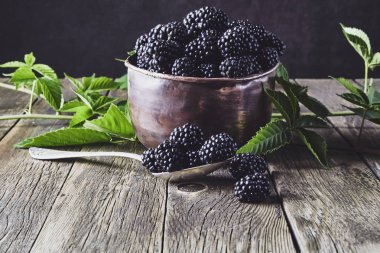 Eski ahşap tahtalar üzerinde eski bakır tabaklarda yaprakları ile Olgun böğürtlen ve koyu bir arka plan üzerinde bir kaşık yanında yalan.