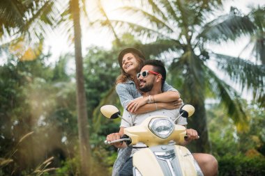 A loving couple travels by scooter through the jungle in Southeast Asia. Smiling and laughing happily clipart