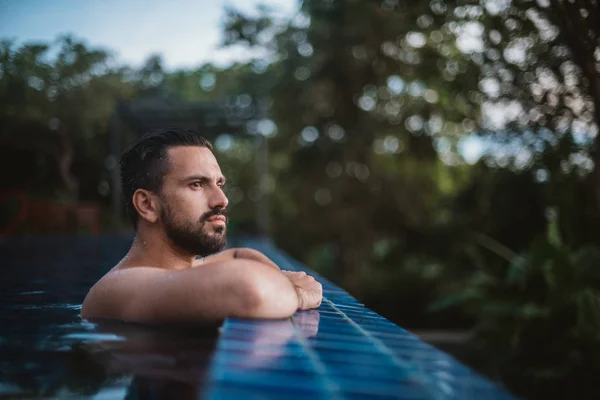 Portrait of a young handsome man in an outdoor pool in the tropics. Attractive guy with a beard at the side of the pool