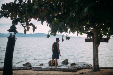 Genç bir adam deniz kıyısı yürüyor. Gün batımında okyanus ufukta güneş gözlüğü yakışıklı adam bakar.