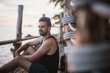 Genç bir adam bir ahşap köprü üzerinde günbatımında okyanusta oturuyor. Yakışıklı adam okyanusun Güneydoğu Asya'da tarafından gün batımını izlemek