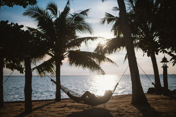 A young man is lying in a hammock at sunset by the ocean. Handsome guy is resting in a hammock among palm trees on the seashore.