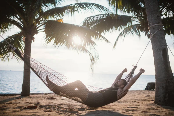 A young man is lying in a hammock at sunset by the ocean. Handsome guy is resting in a hammock among palm trees on the seashore.