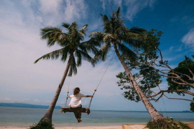 Bir tropik ada plajda palmiye ağaçları arasında bir salıncak üzerinde sallanan bir genç kız. Genç kadın okyanus güneşli bir günde izlerken