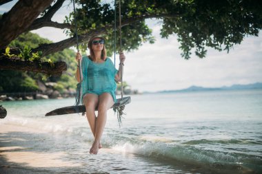 Genç bir kadın deniz kenarında büyük bir ağaç gölgesinde bir salıncak üzerinde sallanıyor. Kız bir tropik ada'nın kıyısındaki beach salıncak üzerinde.