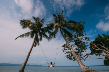 Bir tropik ada plajda palmiye ağaçları arasında bir salıncak üzerinde sallanan bir genç kız. Genç kadın okyanus güneşli bir günde izlerken