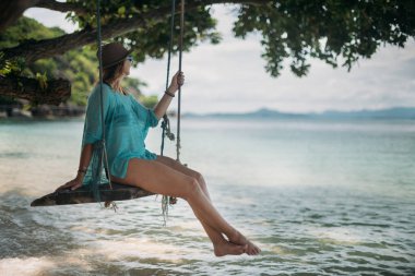 Genç bir kadın deniz kenarında büyük bir ağaç gölgesinde bir salıncak üzerinde sallanıyor. Kız bir tropik ada'nın kıyısındaki beach salıncak üzerinde.