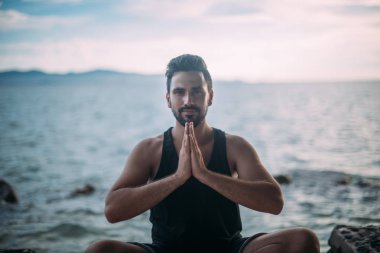 Denizin kenarında yoga yaparken genç adam. Yakışıklı bir adam okyanusun üzerinde günbatımında oturuyor.
