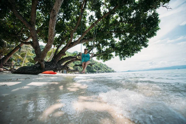 若い女性が海のそばの大きな木の陰にあるブランコに揺れています 熱帯の島の海岸にビーチ スイングの少女 — ストック写真