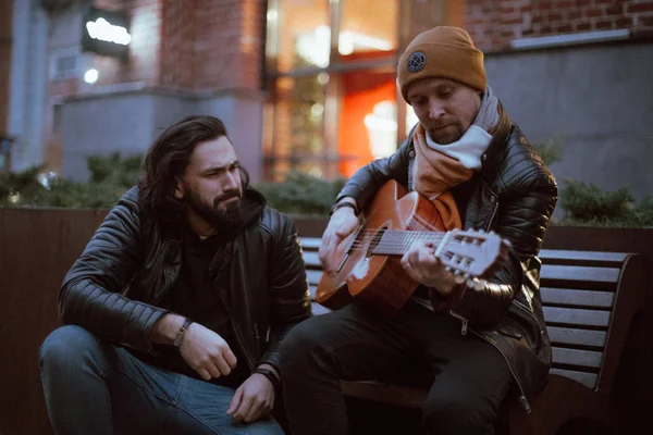 A company of friends on the bench plays the guitar. Young people sit on a bench in the yard with a guitar in the fall