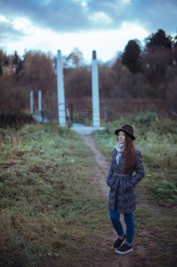 Genç kadın köyde büyük bir asma köprü üzerinde. Kız bir palto ve şapka bir sonbahar bulutlu günde bir ahşap köprü üzerinde yürüyor 