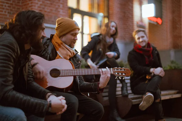 A company of friends on the bench plays the guitar. Young people sit on a bench in the yard with a guitar in the fall