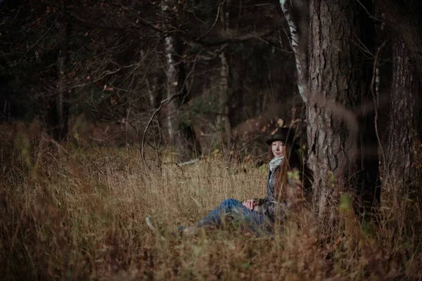 A girl sits alone under a tree in a forest on a sunny day. A young woman with a hat sits thoughtfully on the dry grass in the woods near a tree